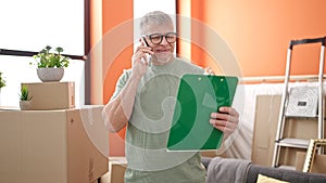 Middle age grey-haired man talking on smartphone reading document at new home