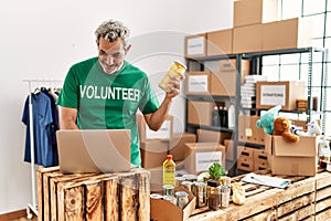 Middle age grey-haired man volunteer using laptop holding bottle with potatoes at charity center