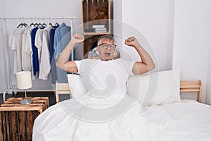 Middle age grey-haired man waking up stretching arms yawning at bedroom