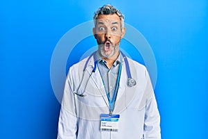 Middle age grey-haired man wearing doctor uniform and stethoscope afraid and shocked with surprise expression, fear and excited