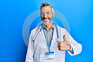 Middle age grey-haired man wearing doctor uniform and stethoscope doing happy thumbs up gesture with hand