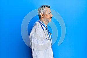 Middle age grey-haired man wearing doctor uniform and stethoscope looking to side, relax profile pose with natural face with