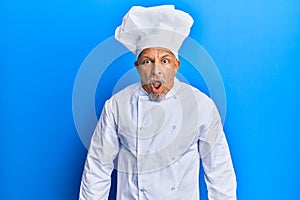 Middle age grey-haired man wearing professional cook uniform and hat afraid and shocked with surprise expression, fear and excited