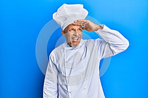 Middle age grey-haired man wearing professional cook uniform and hat very happy and smiling looking far away with hand over head