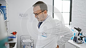 Middle age grey-haired scientist man suffering a backache, toiling over pressing lab work on computer