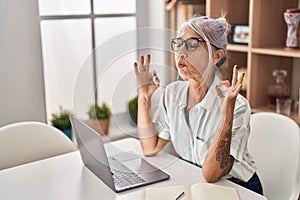 Middle age grey-haired woman doing yoga exercise sitting on table at home