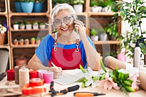 Middle age grey-haired woman florist talking on smartphone writing on notebook at florist