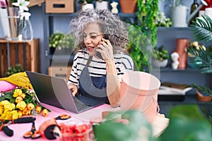 Middle age grey-haired woman florist using laptop talking on smartphone at florist