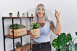 Middle age grey-haired woman holding green plant pot at home smiling positive doing ok sign with hand and fingers