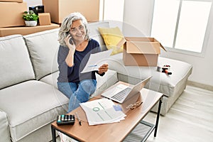 Middle age grey-haired woman sitting on the sofa talking on the smartphone at new home