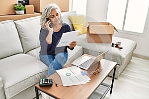 Middle age grey-haired woman sitting on the sofa talking on the smartphone at new home