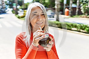 Middle age grey-haired woman smiling happy drinking mate at the city