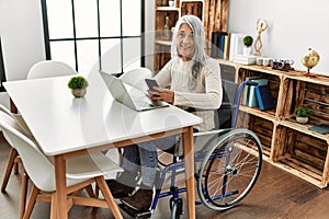 Middle age grey-haired woman using smartphone sitting on wheelchair at home