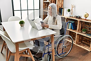 Middle age grey-haired woman using smartphone sitting on wheelchair at home