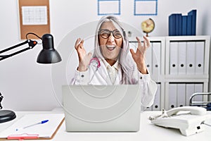 Middle age grey-haired woman wearing doctor uniform working using computer laptop celebrating mad and crazy for success with arms