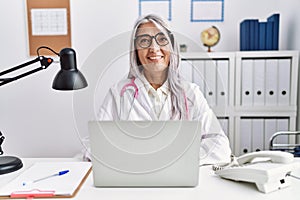 Middle age grey-haired woman wearing doctor uniform working using computer laptop looking positive and happy standing and smiling