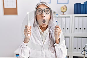 Middle age grey-haired woman working at dentist clinic holding electric teethbrush and toothbrush afraid and shocked with surprise