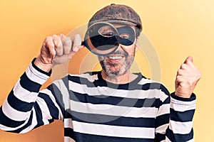 Middle age handsome burglar man searching using magnifying glass over yellow background screaming proud, celebrating victory and
