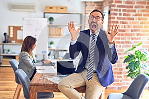 Middle age handsome businessman wearing glasses sitting on desk at the office celebrating mad and crazy for success with arms