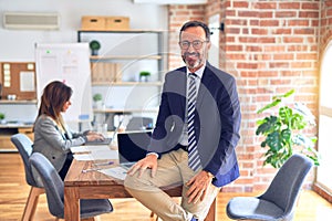 Middle age handsome businessman wearing glasses sitting on desk at the office with a happy and cool smile on face