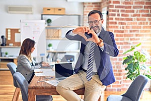 Middle age handsome businessman wearing glasses sitting on desk at the office smiling in love showing heart symbol and shape with