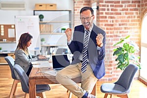 Middle age handsome businessman wearing glasses sitting on desk at the office very happy and excited doing winner gesture with