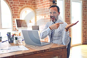 Middle age handsome businessman wearing tie sitting using laptop at the office amazed and smiling to the camera while presenting