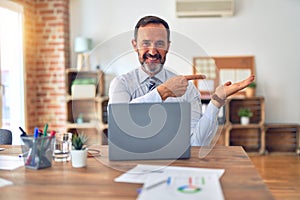 Middle age handsome businessman wearing tie sitting using laptop at the office amazed and smiling to the camera while presenting