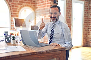 Middle age handsome businessman wearing tie sitting using laptop at the office doing happy thumbs up gesture with hand