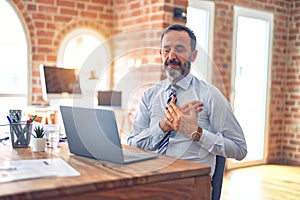 Middle age handsome businessman wearing tie sitting using laptop at the office smiling with hands on chest with closed eyes and
