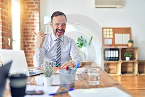 Middle age handsome businessman wearing tie sitting using laptop at the office smiling with happy face looking and pointing to the