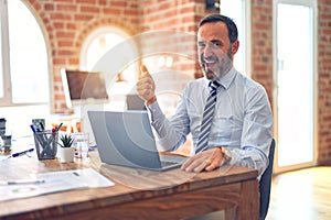 Middle age handsome businessman wearing tie sitting using laptop at the office smiling with happy face looking and pointing to the