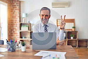 Middle age handsome businessman wearing tie sitting using laptop at the office smiling with happy face winking at the camera doing