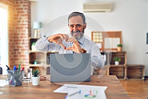 Middle age handsome businessman wearing tie sitting using laptop at the office smiling in love showing heart symbol and shape with