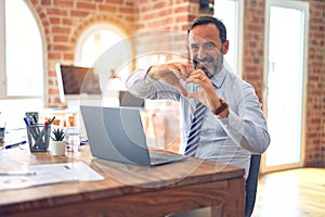 Middle age handsome businessman wearing tie sitting using laptop at the office smiling in love showing heart symbol and shape with
