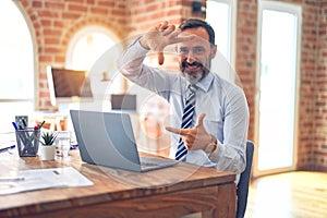 Middle age handsome businessman wearing tie sitting using laptop at the office smiling making frame with hands and fingers with