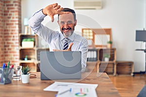 Middle age handsome businessman wearing tie sitting using laptop at the office smiling making frame with hands and fingers with