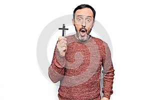 Middle age handsome catholic man praying holding christian cross over isolated background scared and amazed with open mouth for