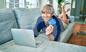 Middle age handsome caucasian man relaxing at home doing online shopping using credit card on computer laptop