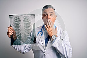 Middle age handsome grey-haired doctor man holding chest xray over white background cover mouth with hand shocked with shame for