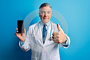 Middle age handsome grey-haired doctor man holding smartphone showing screen happy with big smile doing ok sign, thumb up with