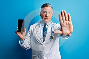 Middle age handsome grey-haired doctor man holding smartphone showing screen with open hand doing stop sign with serious and