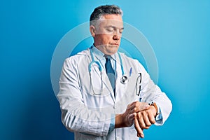 Middle age handsome grey-haired doctor man wearing coat and blue stethoscope Checking the time on wrist watch, relaxed and