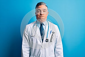 Middle age handsome grey-haired doctor man wearing coat and blue stethoscope puffing cheeks with funny face