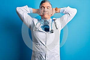 Middle age handsome grey-haired doctor man wearing coat and blue stethoscope relaxing and stretching, arms and hands behind head