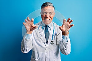 Middle age handsome grey-haired doctor man wearing coat and blue stethoscope smiling funny doing claw gesture as cat, aggressive