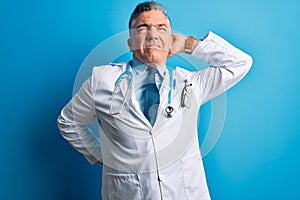 Middle age handsome grey-haired doctor man wearing coat and blue stethoscope Suffering of neck ache injury, touching neck with