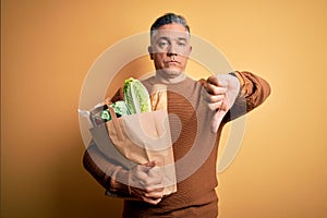 Middle age handsome grey-haired man holding paper bag with food over yellow background with angry face, negative sign showing