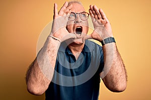 Middle age handsome hoary man wearing casual polo and glasses over yellow background Shouting angry out loud with hands over mouth