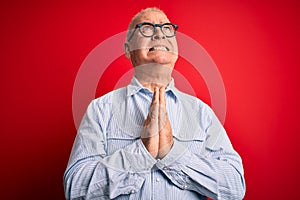 Middle age handsome hoary man wearing casual striped shirt and glasses over red background begging and praying with hands together